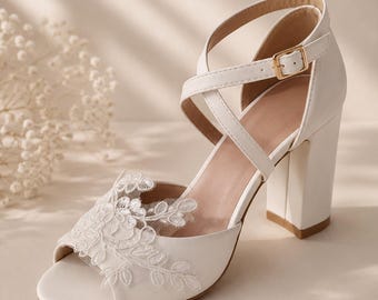 May include: A white open-toe block heel shoe featuring a delicate floral lace overlay on the front strap and crisscross ankle straps with a gold-toned metal buckle closure. The shoe has a sturdy, thick heel and a smooth, solid white finish.