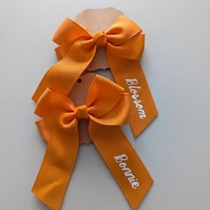 May include: Two bright orange grosgrain ribbon hair bows attached to brown cardstock tags. Each bow features long, trailing tails with white script text printed on the ends. One bow displays the name Blossom, and the other displays the name Bonnie.