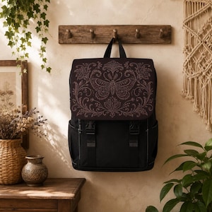 Op de afbeelding: Een zwarte rugzak met een bovenflap voorzien van een gedetailleerd, ingewikkeld motief van een nachtvlinder en bloemen in roségoud. De tas heeft twee verticale zwarte banden met kunststof gespen, zijvakken en een handvat aan de bovenzijde.