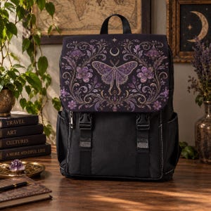 Peut inclure: Un sac à dos noir doté d'un rabat orné d'une illustration de papillon de nuit violet, de motifs floraux et d'un symbole de croissant de lune. Le sac comprend deux fermetures à boucle en plastique noir, une poignée et des poches latérales.