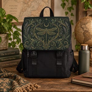 Peut inclure: Un sac à dos noir doté d'un rabat vert foncé orné d'une illustration de libellule dorée, de volutes botaniques complexes et d'un symbole de croissant de lune. Le sac comprend deux fermetures à boucle en plastique noir et des bretelles réglables.