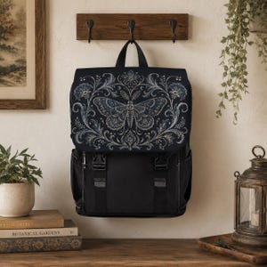 Peut inclure: Un sac à dos noir doté d'un rabat orné d'un motif complexe de papillon de nuit et de fleurs en blanc et bleu clair. Le sac comprend deux fermetures à boucle en plastique noir sur le devant, une poignée supérieure et des poches latérales.