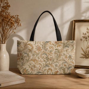 May include: A tote bag featuring a cream-colored base with an intricate, repeating floral and scrollwork pattern in muted shades of gold, sage green, and dusty blue. The bag has two solid black fabric handles attached to the top edge.
