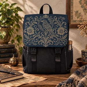 May include: A black backpack featuring a dark blue flap with a gold-toned graphic print of a raven centred within a circular frame, surrounded by intricate floral and vine patterns. The bag includes two vertical black straps with buckle closures.