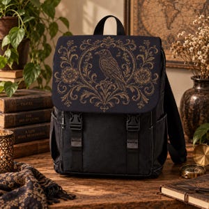 Può includere: Uno zaino nero con una patta caratterizzata da una stampa grafica color oro raffigurante un corvo al centro di una cornice floreale decorata. La borsa include due cinghie verticali con fibbie in plastica nera, tasche laterali e una maniglia.