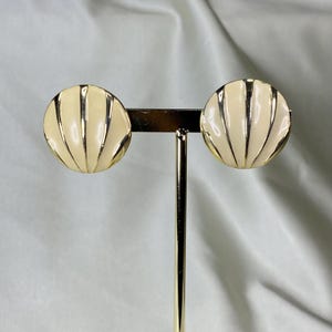 Op de afbeelding: Een paar ronde, crèmekleurige emaille oorbellen in de vorm van schelpen. Elke oorbel is voorzien van een reeks verticale, verhoogde goudkleurige metalen ribbels die vanuit het midden naar de buitenrand uitstralen, wat een waaiervormig patroon creëert.