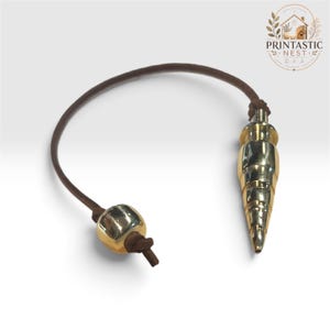 May include: A gold-toned metal pendulum with a pointed, tiered design attached to a brown leather cord. A single, smooth, spherical gold-toned metal bead is threaded onto the cord, positioned near the end of the leather strap.