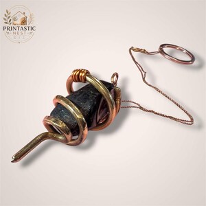 May include: A dark, raw, faceted crystal pendant wrapped in a thick, spiraling copper wire cage. The pendant is attached to a thin, delicate copper chain that features a circular metal ring at the end, creating a distinct, metallic, and earthy aesthetic.