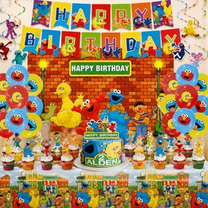 May include: A Sesame Street themed birthday party set featuring a Happy Birthday banner, red, yellow, and blue balloons, a blue cake with the text Happy Birthday Alden 2, and a tablecloth decorated with Sesame Street character illustrations.