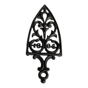 May include: A black cast iron trivet shaped like a flat iron, featuring an intricate openwork scroll design and the embossed numbers 1884 centered within the metal frame. The object has a pointed top and a small circular loop at the base.