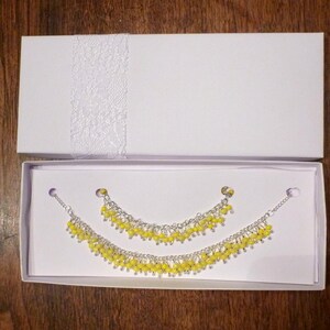May include: A set of two yellow beaded bracelets in a white box. The bracelets are made of small, round beads and are connected by a silver chain.