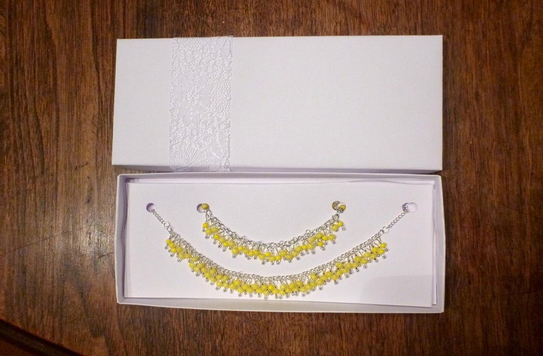 May include: A set of two yellow beaded bracelets in a white box. The bracelets are made of small, round beads and are connected by a silver chain.