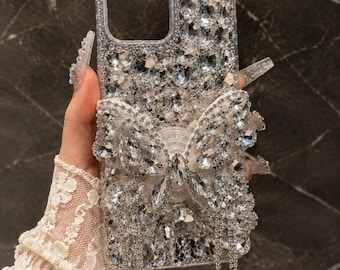 Peut inclure: Une coque de téléphone transparente recouverte de strass argentés étincelants de différentes tailles, avec un grand papillon central orné de cristaux et deux longues chaînes à franges en cristal pendantes sous le papillon.