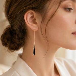 May include: A long gold-tone drop earring featuring a thin chain attached to a small stud at the top and a sleek, elongated teardrop-shaped pendant at the bottom. The pendant has a polished gold-tone border surrounding a smooth black center inlay.