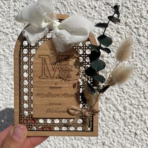 Peut inclure: Une plaque en bois arquée avec la lettre M gravée au laser, des motifs floraux et le texte "comme Meilleure Atsem comme Mélanie". Elle présente une bordure en cannage, un nœud en tissu blanc et des tiges d'eucalyptus et d'herbes séchées.