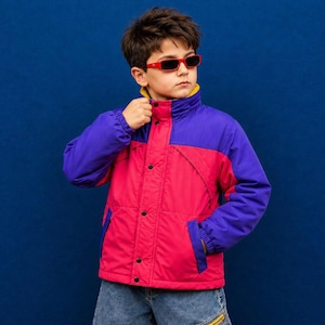 May include: A color-blocked jacket featuring a bright pink torso and purple sleeves with a high collar. The jacket has a front zipper closure with black snap buttons, a yellow inner lining, and is worn with red-framed sunglasses and blue denim jeans.