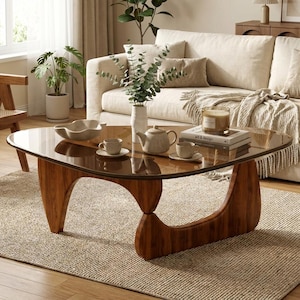 Puede incluir: Una mesa de centro con tapa de cristal marrón oscuro y base de madera curva. Sobre la mesa hay una tetera de cerámica blanca, dos tazas a juego con platillos, un cuenco blanco y dos libros con el texto "art & design" en la portada.