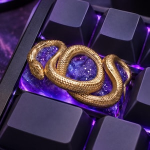 May include: A custom mechanical keyboard keycap featuring a coiled gold-colored snake resting on a translucent purple resin base with embedded glitter, surrounded by matte black keys with purple under-key lighting.