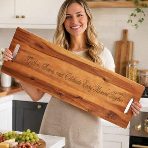 May include: A rectangular wooden serving tray with a natural wood grain finish and two silver-toned metal handles on the ends. The center of the tray is engraved with the text, Gather, Savor, and Celebrate Every Moment Together, in a cursive font.