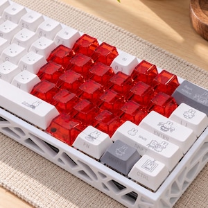 Könnte beinhalten: Eine mechanische Tastatur mit einem weißen Rahmen in Gitteroptik, weißen Tastenkappen mit schwarzer Beschriftung und Miffy-Illustrationen sowie einem zentralen Block aus durchscheinenden roten Tastenkappen. Inklusive Tasten wie Enter, Shift und Strg.