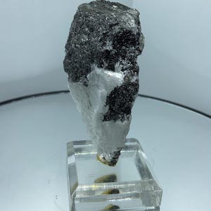 May include: A raw mineral specimen featuring a dark, textured, crystalline upper section contrasted with a smooth, opaque white base. The specimen is mounted vertically on a clear, square acrylic block stand, showcasing its natural, irregular shape.