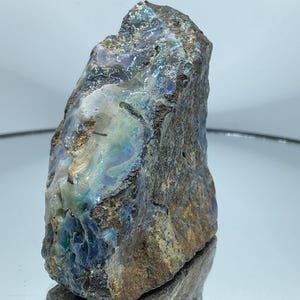 Puede incluir: Un espécimen de ópalo boulder en bruto que presenta una matriz rugosa de color marrón oscuro con destellos iridiscentes de luz azul, verde y violeta en su superficie irregular y texturizada. La piedra muestra una forma natural y dentada.