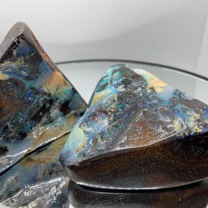 May include: Two polished boulder opal specimens with irregular triangular shapes. The stones feature a dark brown ironstone matrix with vibrant flashes of blue, green, and cream-coloured opal veins. The surfaces are smooth and reflective.