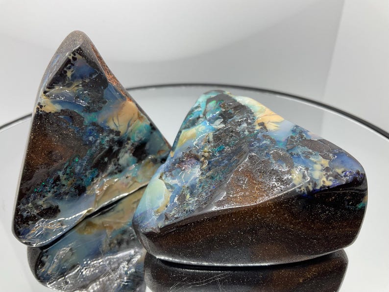 May include: Two polished boulder opal specimens with irregular triangular shapes. The stones feature a dark brown ironstone matrix with vibrant flashes of blue, green, and cream-coloured opal veins. The surfaces are smooth and reflective.