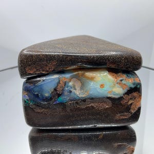 May include: A raw boulder opal specimen featuring a dark brown ironstone matrix with a prominent horizontal band of iridescent blue, green, and orange opal play-of-colour. The stone has a smooth, polished top surface and a rough, natural texture.