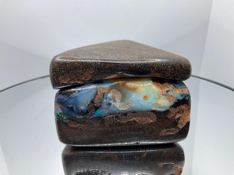 May include: A raw boulder opal specimen featuring a dark brown ironstone matrix with a prominent horizontal band of iridescent blue, green, and orange opal play-of-colour. The stone has a smooth, polished top surface and a rough, natural texture.