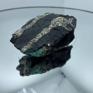 以下が含まれることがあります： 暗灰色から黒色の粗い鉱物標本で、表面には金属光沢のある金色の黄鉄鉱の目立つギザギザした脈が走り、石の下端近くには鮮やかな緑色の鉱物素材がはっきりと見えています。