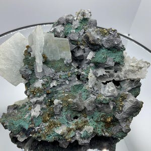 May include: A mineral specimen featuring a cluster of translucent white calcite crystals, dark grey galena, vibrant green malachite, and small metallic gold-colored pyrite inclusions. The geological formation displays a complex, jagged texture.