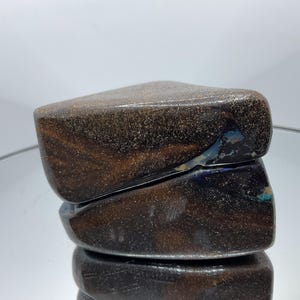 May include: A polished boulder opal gemstone with a dark brown, ironstone matrix featuring fine, glittering gold-toned flecks and a distinct, iridescent blue flash visible along the angled, smooth surface of the stone.