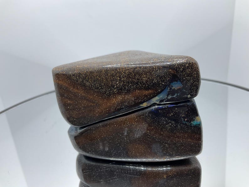 May include: A polished boulder opal gemstone with a dark brown, ironstone matrix featuring fine, glittering gold-toned flecks and a distinct, iridescent blue flash visible along the angled, smooth surface of the stone.