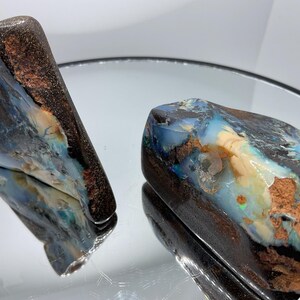 May include: Two raw boulder opal specimens featuring dark brown ironstone matrix with vibrant flashes of blue, green, and white iridescent colour play. The stones have irregular, organic shapes with polished surfaces that reflect light.