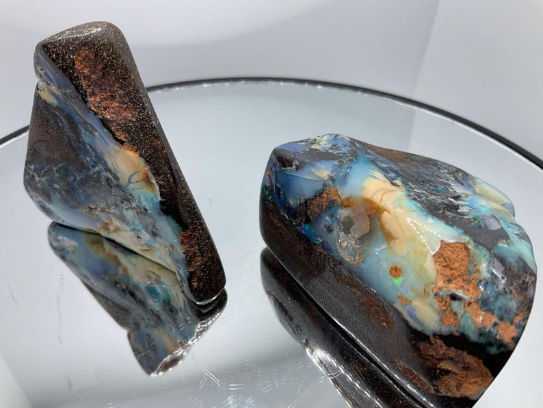 May include: Two raw boulder opal specimens featuring dark brown ironstone matrix with vibrant flashes of blue, green, and white iridescent colour play. The stones have irregular, organic shapes with polished surfaces that reflect light.