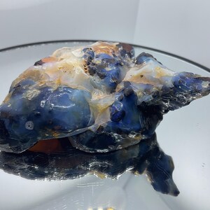 May include: A raw, unpolished opal specimen featuring deep blue and translucent white coloration with patches of amber and brown. The stone has an irregular, organic shape with a glossy, reflective surface texture and visible internal inclusions.