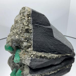 Może przedstawiać: Okaz minerału z ciemnoszarą, warstwową matrycą skalną, w której osadzone są metaliczne, złote kryształy pirytu oraz kilka żywo zielonych, sześciokątnych kryształów szmaragdu wystających z boku formacji skalnej.
