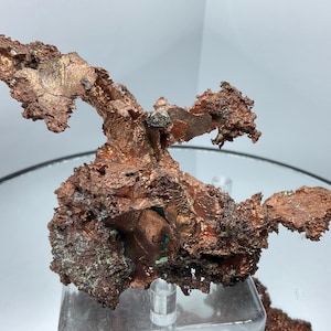 May include: A raw, metallic copper mineral specimen featuring a complex, branching crystalline structure with a reddish-brown, oxidized surface. The formation displays jagged, porous textures and metallic luster, supported by a clear acrylic stand.