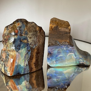 May include: Two raw boulder opal specimens featuring rough, earthy brown rock surfaces contrasted with polished, iridescent sections displaying vibrant flashes of blue, green, and orange light, reflecting clearly on a smooth, circular surface.