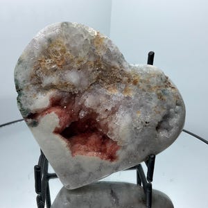 May include: A heart-shaped stone specimen featuring a central geode cavity filled with deep red and translucent white crystalline structures. The outer surface is a smooth, pale grey mineral with patches of tan and dark green inclusions.