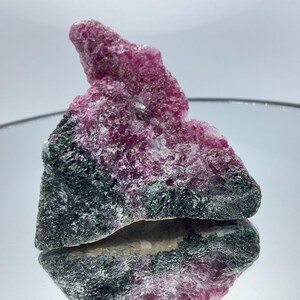 May include: A raw mineral specimen featuring a vibrant magenta-pink crystalline cluster atop a dark, textured charcoal-gray base. The stone has an irregular, jagged shape with a sparkling, multifaceted surface that reflects light across its crystalline structure.