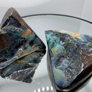 May include: Two triangular boulder opal specimens featuring a dark brown ironstone matrix with iridescent flashes of blue, green, and cream. The polished surfaces exhibit a glossy, reflective texture with natural mineral inclusions and veins.