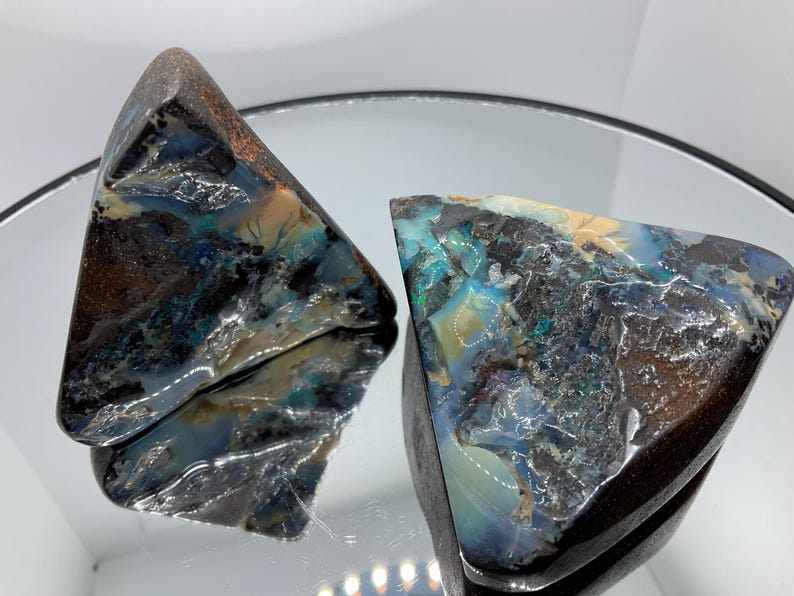 May include: Two triangular boulder opal specimens featuring a dark brown ironstone matrix with iridescent flashes of blue, green, and cream. The polished surfaces exhibit a glossy, reflective texture with natural mineral inclusions and veins.