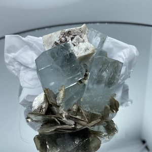 May include: A cluster of pale blue, translucent hexagonal aquamarine crystals attached to a matrix of white feldspar and brownish-grey mica flakes. The mineral specimen features sharp, well-defined crystal terminations and a natural, raw texture.