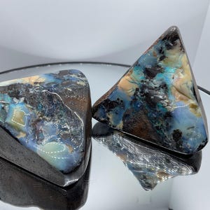 Puede incluir: Dos especímenes de ópalo boulder de forma triangular con una matriz de piedra de hierro marrón oscuro y vetas iridiscentes de color azul, verde y crema. Las superficies están pulidas con un alto brillo, mostrando variaciones de color.