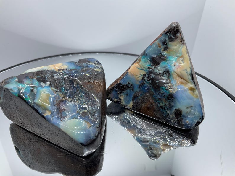 May include: Two triangular-shaped boulder opal specimens featuring a dark brown ironstone matrix with vibrant flashes of iridescent blue, green, and cream-coloured opal veins. The surfaces are polished to a high gloss, showing natural colour variations.