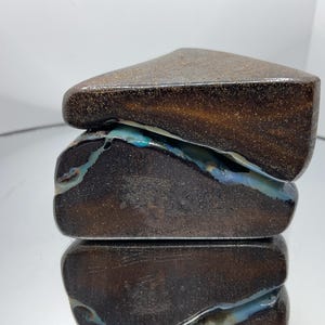 May include: A polished boulder opal specimen featuring a dark brown, ironstone matrix with a prominent, irregular vein of iridescent blue and green opal running horizontally through the centre of the stone, showcasing a smooth, rounded surface.