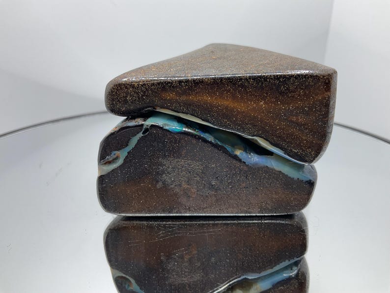 May include: A polished boulder opal specimen featuring a dark brown, ironstone matrix with a prominent, irregular vein of iridescent blue and green opal running horizontally through the centre of the stone, showcasing a smooth, rounded surface.