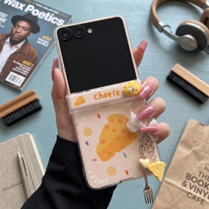 May include: A clear foldable smartphone case featuring a cheese-themed design with yellow Swiss cheese illustrations, the word cheese, and a small 3D cheese charm. A silver-toned metal charm chain with a tiny fork and cheese wedge is attached.
